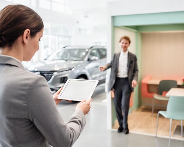 自動車ディーラーのショールームでスタッフがタブレットを確認し、商談ブースへ来店客を案内する様子